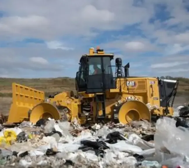 A yellow CAT bulldozer moves through a landfill, pushing aside heaps of trash under a cloudy sky
